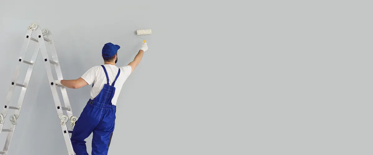 Professional painter in blue overalls using a roller to paint a wall light gray.