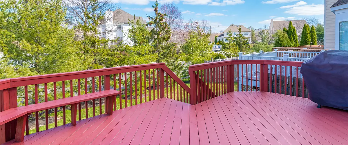 Spacious red-painted backyard deck with a scenic view of trees and neighboring houses under a bright blue sky.