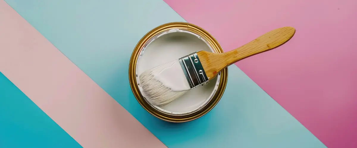 Top view of an open can of white paint with a paintbrush, set on a pastel blue and pink striped background.