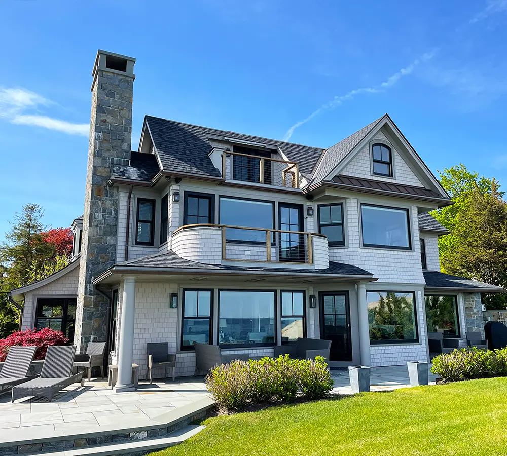 Luxury waterfront home with stone chimney, manicured lawn, and large glass windows.