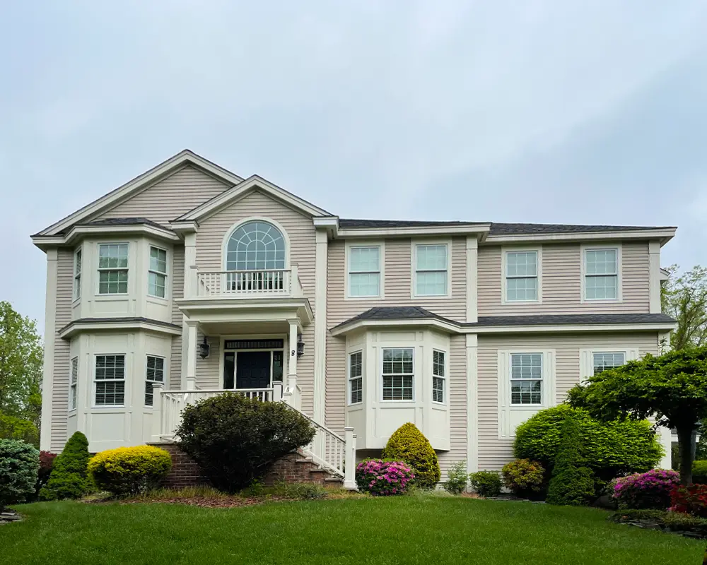 Elegant beige home with landscaped front yard and bright windows under overcast sky.