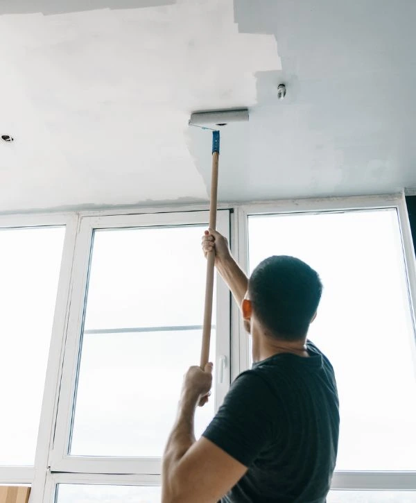 A professional painter using a roller brush on an extended pole to paint a white ceiling, with large windows in the background, representing an interior painting company.