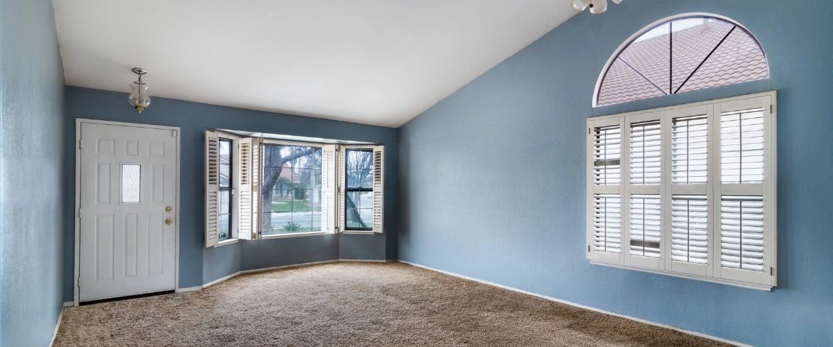 A freshly painted living room with light blue walls, white trim, and large windows featuring plantation shutters, showcasing professional interior painting services.