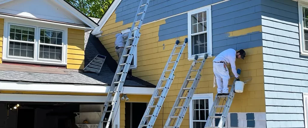 House exterior painting in progress with painters on ladders applying blue paint over yellow siding for a fresh, updated look.