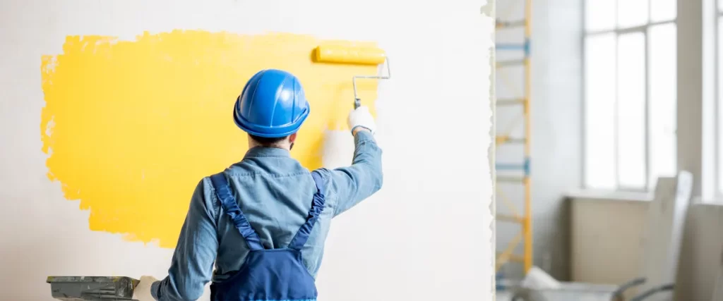 Worker wearing a blue hard hat using a roller brush to apply yellow paint on a wall.