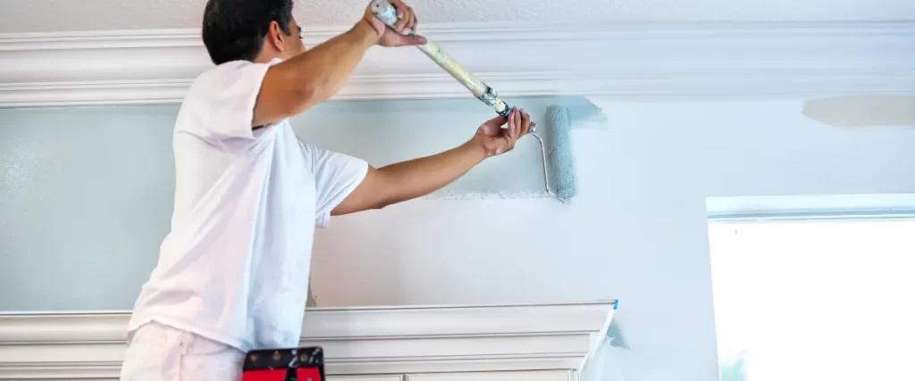 Painter in a white shirt using a small brush to paint a wall near the ceiling.