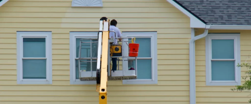 Painter using a hydraulic lift to paint the exterior of a yellow house.