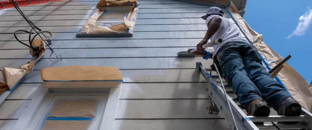 Two workers painting house siding using ladders and rollers.