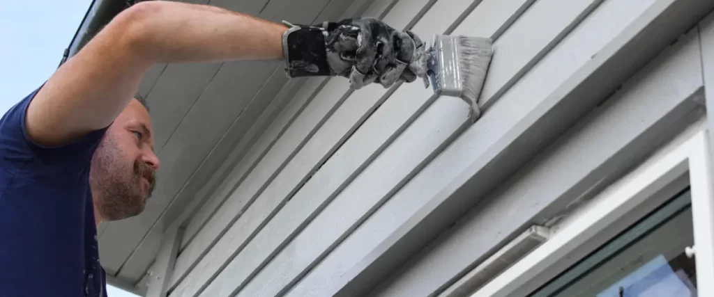 Close-up of a gloved hand painting exterior siding with a brush.